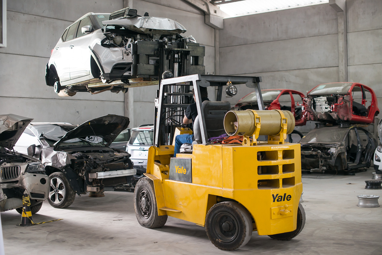 empilhadeira levantando sucata de um automóvel com um ferro-velho ao fundo